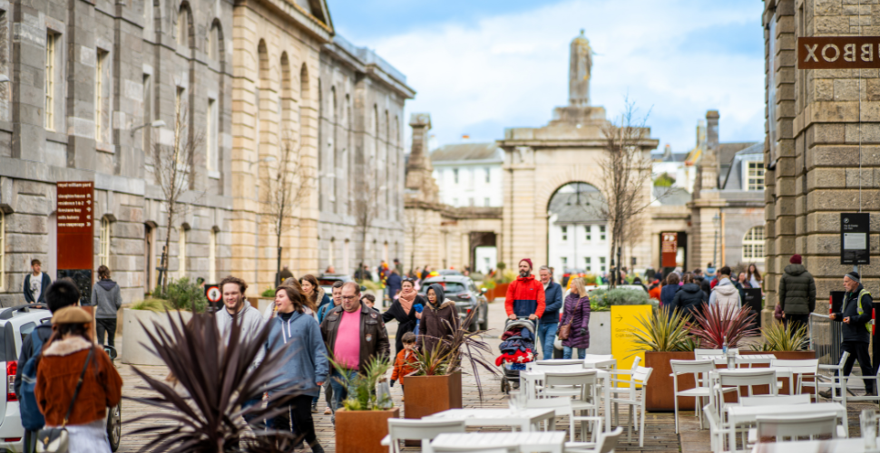 Busy pedestrian street with historic stone buildings, outdoor café seating, and people walking and socialising on a bright day.